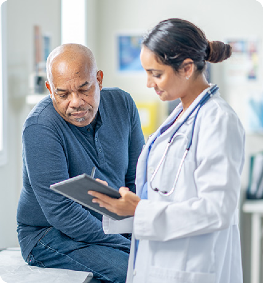 A doctor in a white coat reviews information on a tablet with a man seated on an exam table in a medical office.