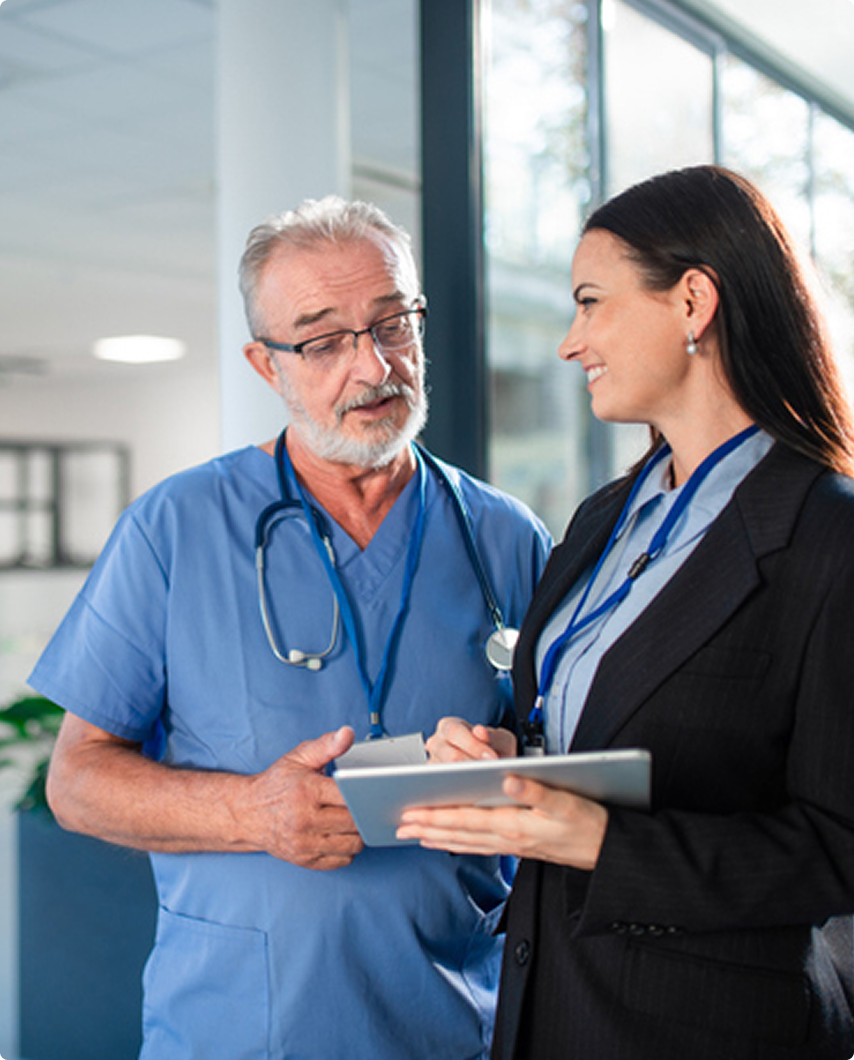 A male doctor in scrubs talks with a woman in business attire holding a tablet. They are standing in a modern medical facility.